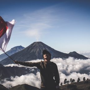 man wearing black crew neck sweater holding white and red flag standing near mountain under blue and white sky